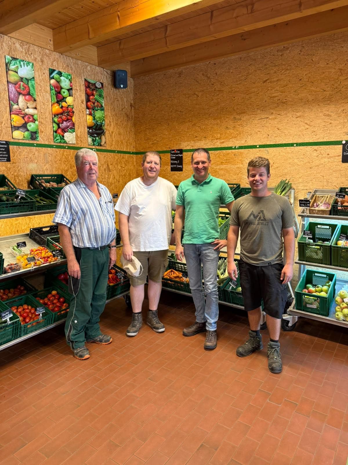 Vier Männer stehen im Supermarkt vor Obst- und Gemüseständen, lächeln in die Kamera, im Hintergrund bunte Obstbilder an der Wand.