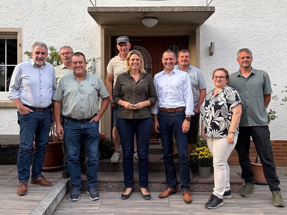 Gruppenfoto mit acht Personen vor einem weißen Haus, alle lächelnd und in Business-Casual Kleidung, bei Tageslicht aufgenommen.