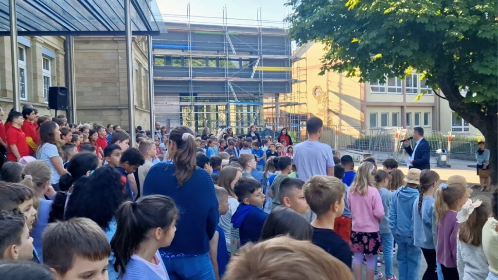 Viele Kinder und Jugendliche versammeln sich im Freien bei einer Schulveranstaltung, um einem Redner zuzuhören, mit Bäumen und Gebäuden im Hintergrund.