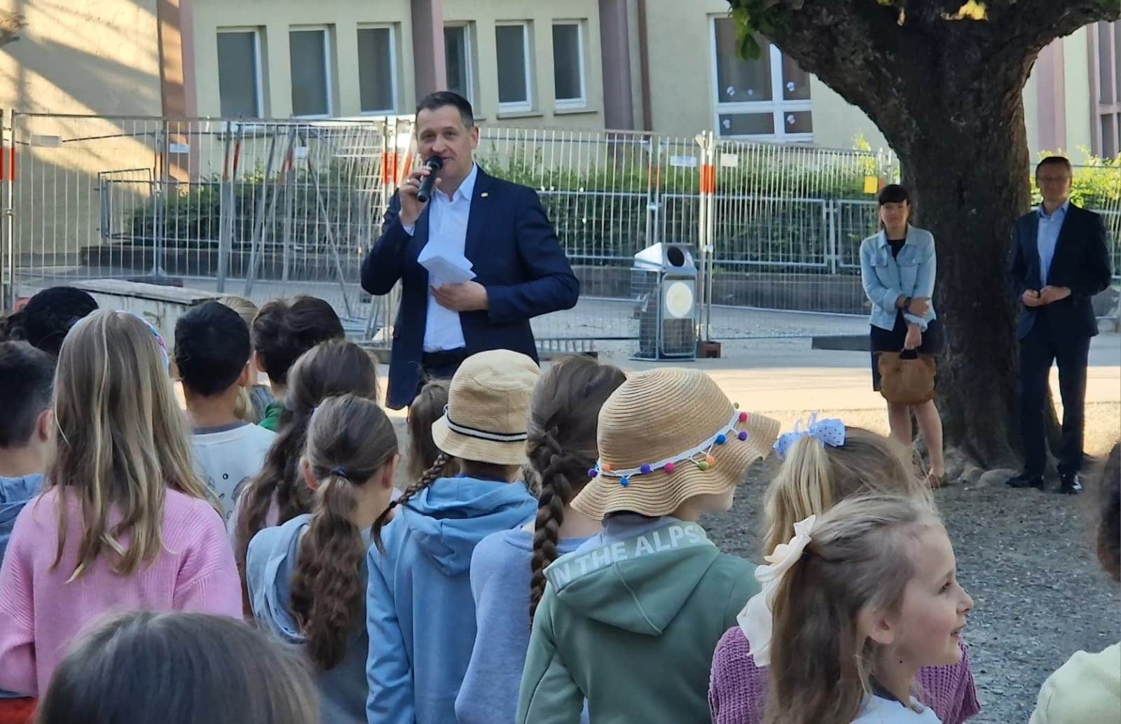 Christoph Stetter spricht bei einer Veranstaltung vor einer Gruppe von Kindern und Jugendlichen im Freien, mit einem Baum und Gebäuden im Hintergrund.