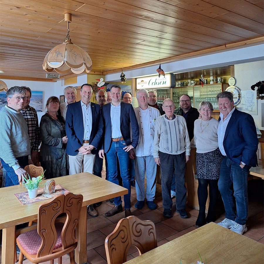 Der Bild zeigt eine Gruppe von Menschen in einem gemütlichen Raum, vermutlich bei einem Treffen oder einer Feier, mit freundlicher Atmosphäre und traditioneller Einrichtung.