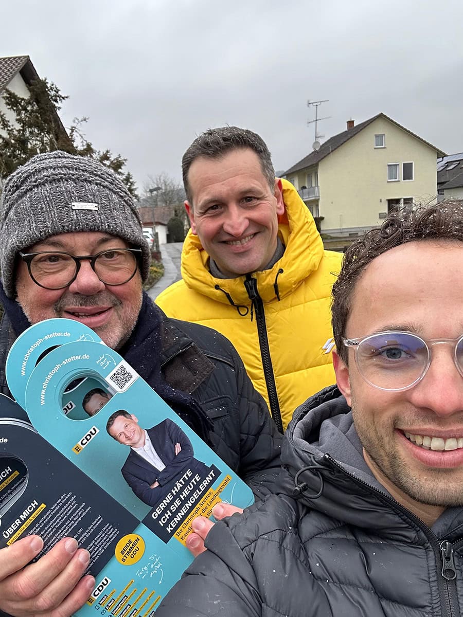 Attraktives Gruppenfoto von drei Männern im Freien bei bewölktem Himmel, einer hält eine Wahlkampf-Flyer in der Hand, alle lächeln freundlich, im Hintergrund moderne Häuser in einer ruhigen Wohngegend.
