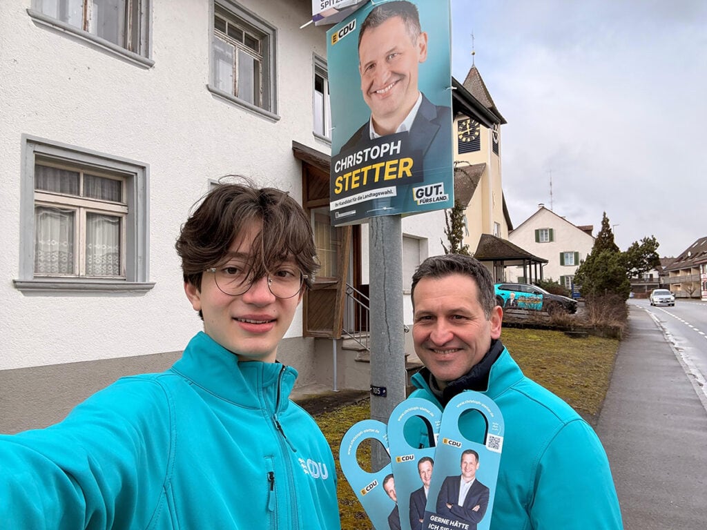 Christoph Stetter bei einem Wahlkampfauftritt, neben einem Wahlplakat mit seinem Bild und Namen, beide tragen blaue Jacken, in einer kleinen Stadt mit Wohnhäusern und einer Kirche im Hintergrund.