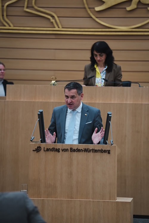 Christoph Stetter spricht im Landtag von Baden-Württemberg während einer Debatte, im Hintergrund eine Frau am Rednerpult.