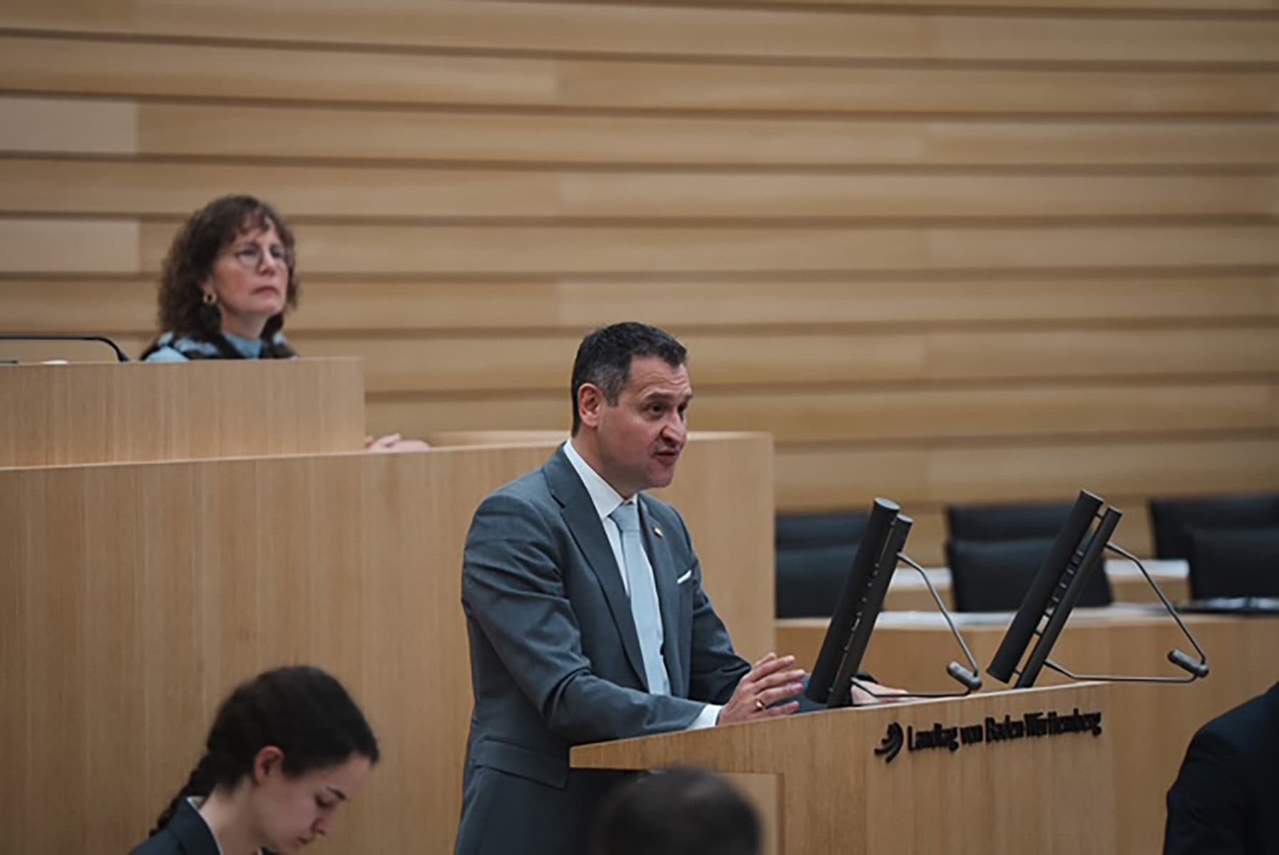 Christoph Stetter spricht im Landtag vor, während eine Frau im Hintergrund zuhört, in einem modernen Sitzungssaal mit Holzvertäfelung.