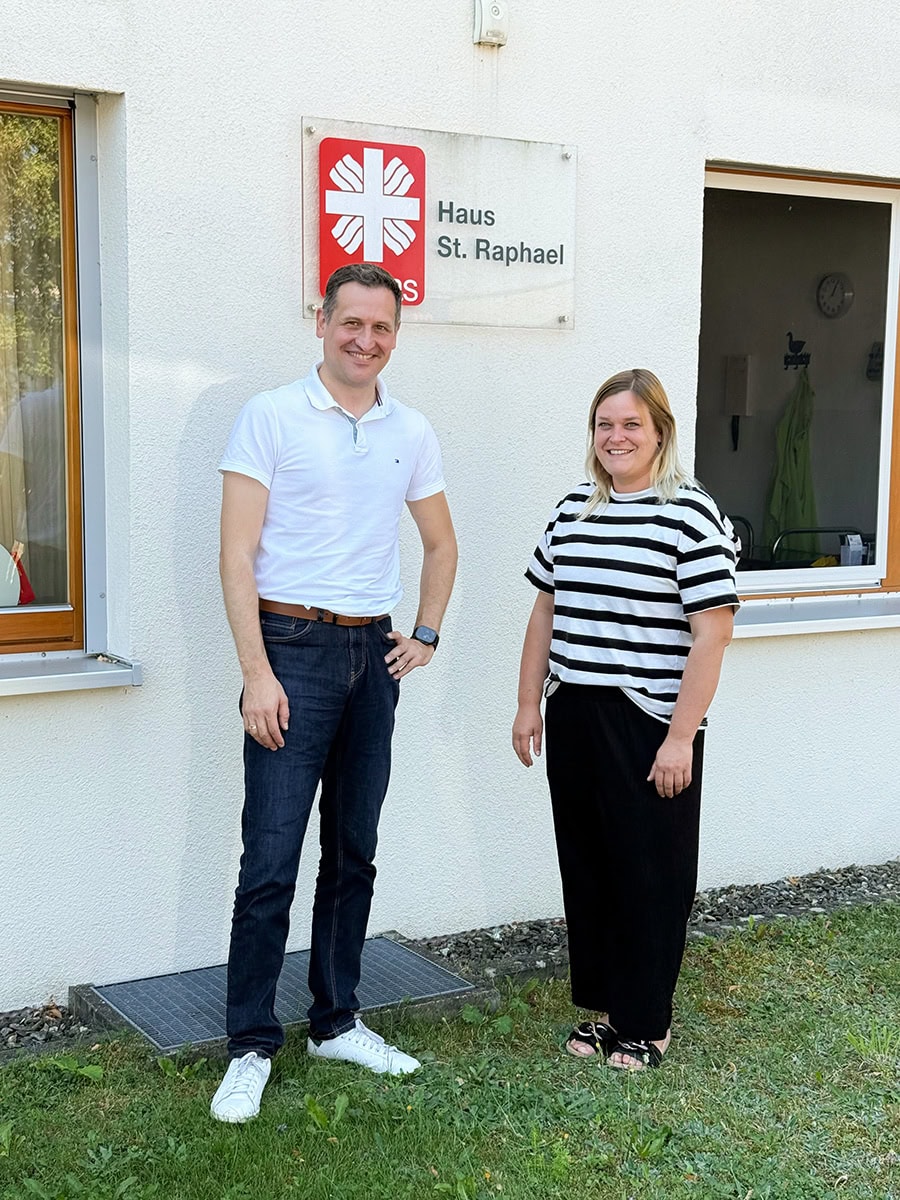 Zwei Personen stehen vor dem Haus St. Raphael, das eine weiße Fassade und ein Schild mit einem roten Kreuz zeigt, im Rahmen des Perspektivwechsels.