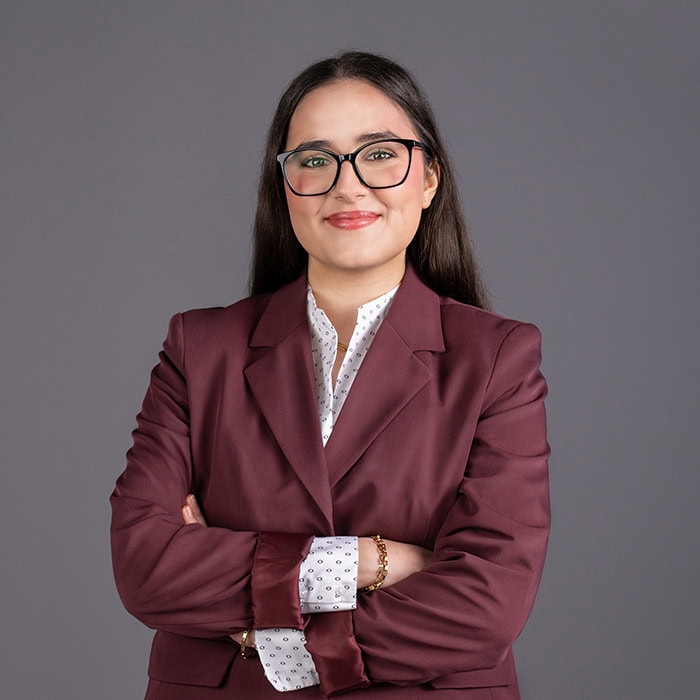 Porträt einer professionellen Frau in Brille und Blazer, die selbstbewusst lächelnd die Arme verschränkt, vor neutralem grauem Hintergrund.