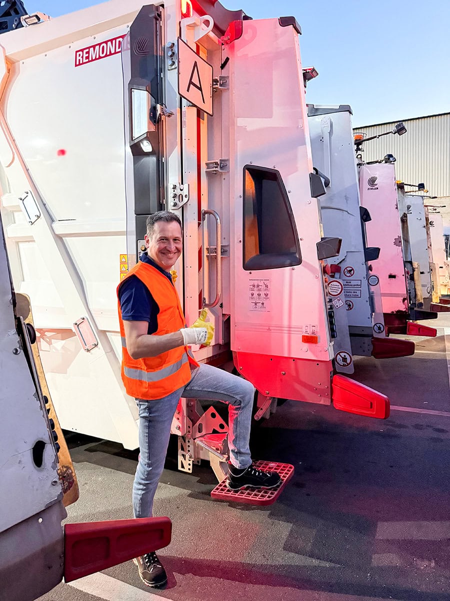Christoph Stetter, lachend und in Sicherheitsweste, steht vor Recyclingmaschinen im Freien, symbolisiert nachhaltiges Arbeiten und Umweltbewusstsein.