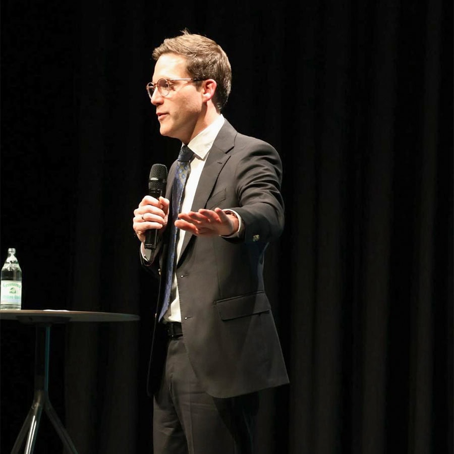 Der Mann in Anzug hält ein Mikrofon und spricht auf einer Bühne mit schwarzem Vorhang im Hintergrund, vermutlich bei einer Präsentation oder einem Vortrag.